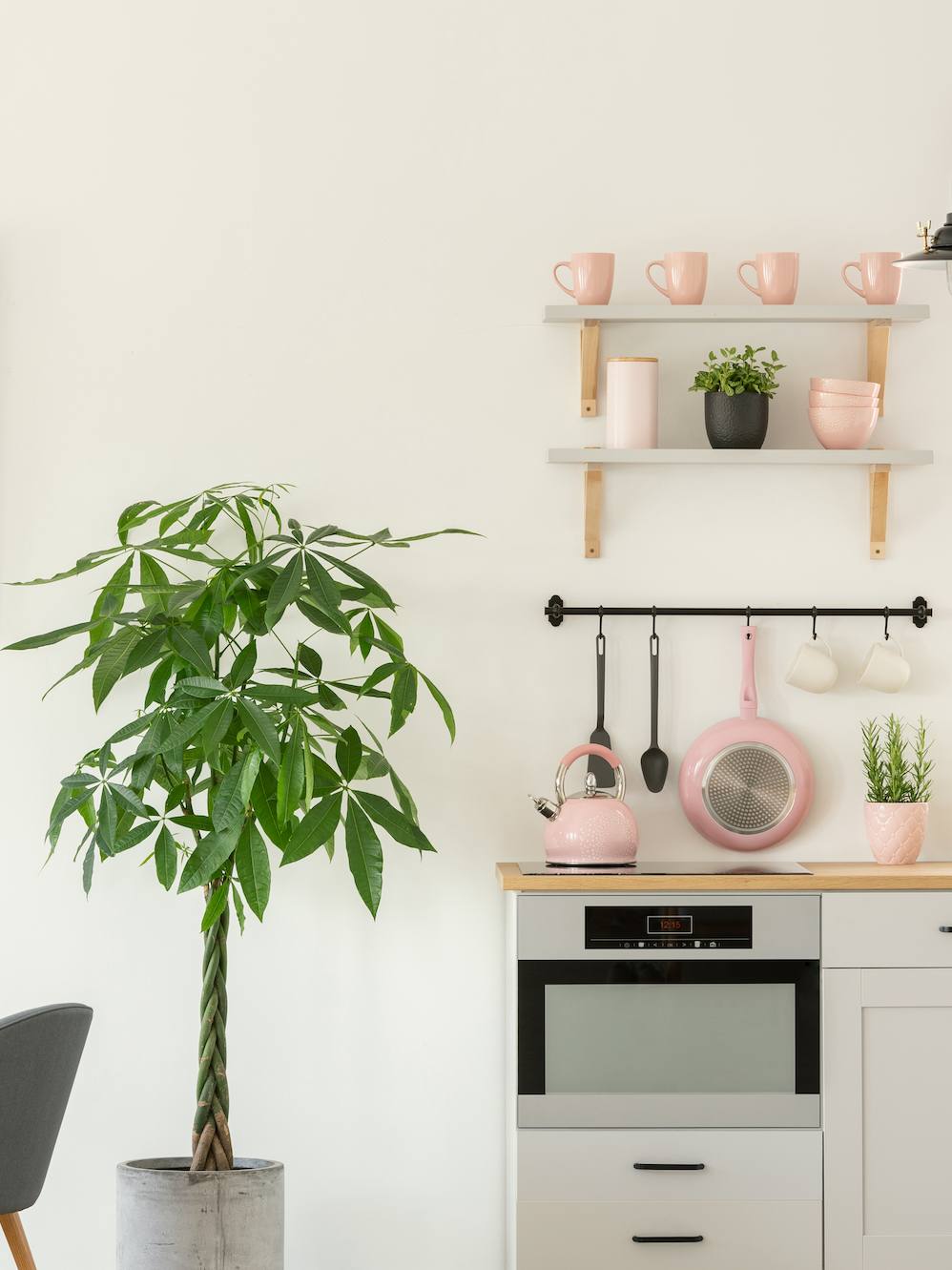 Fresh plant standing next to wooden dining table with chairs in real photo of bright kitchen interior with pastel pink utensils and simple poster on wall